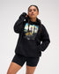 Female model wearing the RUDIS Divine Struggle fleece hoodie in Black with a relaxed fit, drawstring hood, kangaroo pocket and colorful front artwork.