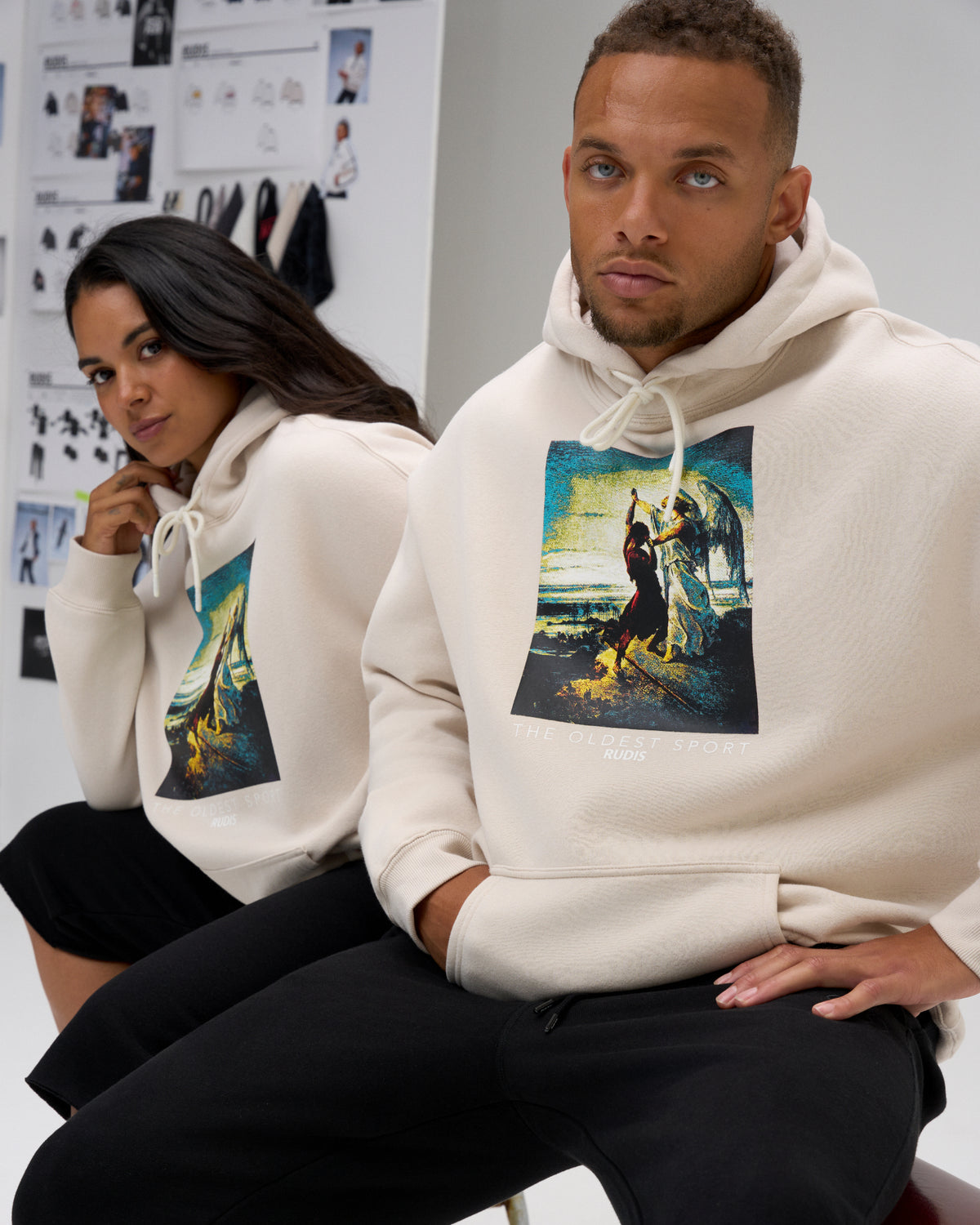 Male and female models seated in RUDIS Divine Struggle fleece hoodies in Bone, highlighting the wrestling-inspired artwork and relaxed comfort fit.