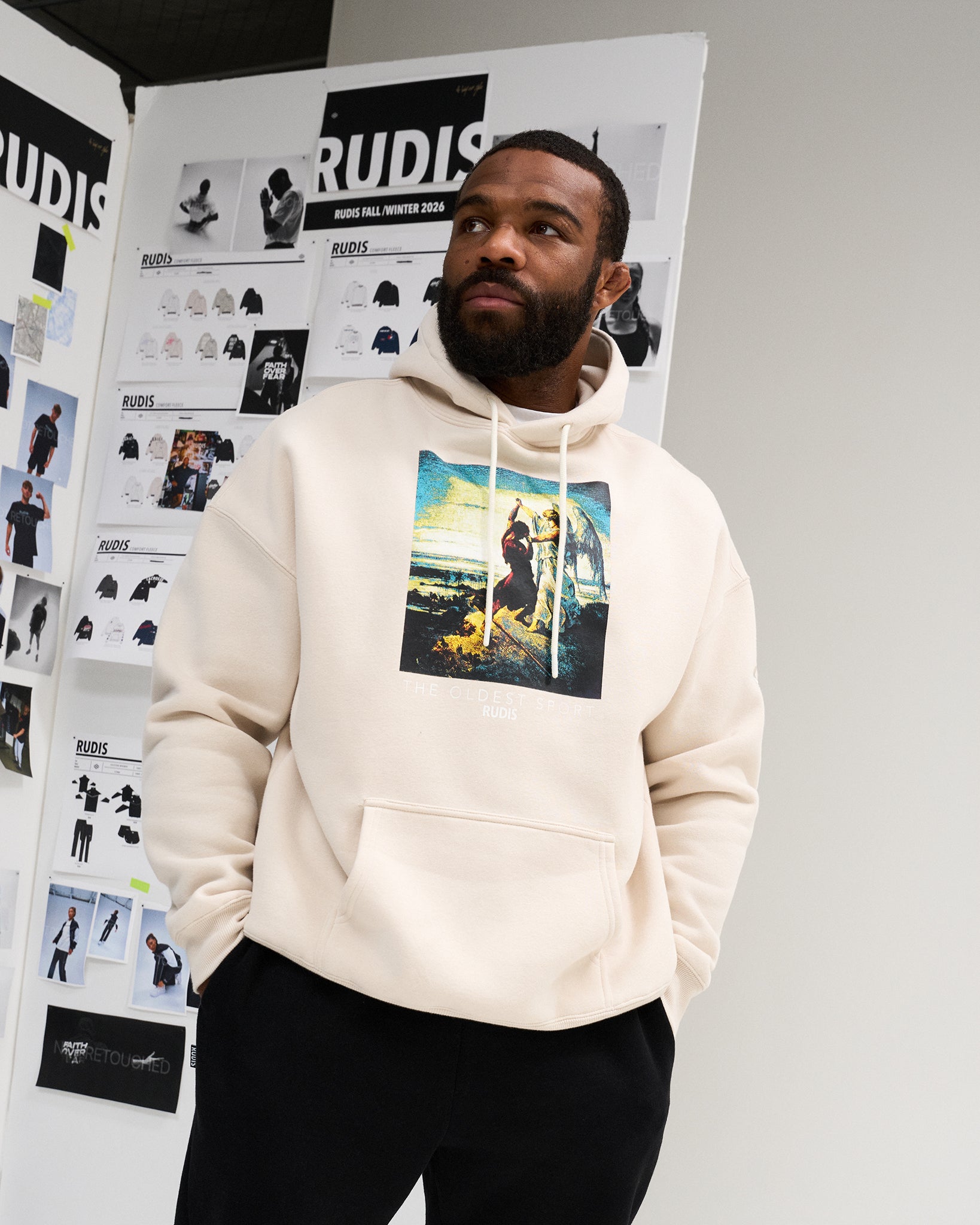 Male model wearing the RUDIS Divine Struggle Relaxed Comfort Fleece Hoodie in Bone, featuring a colorful The Oldest Sport chest graphic and kangaroo pocket.