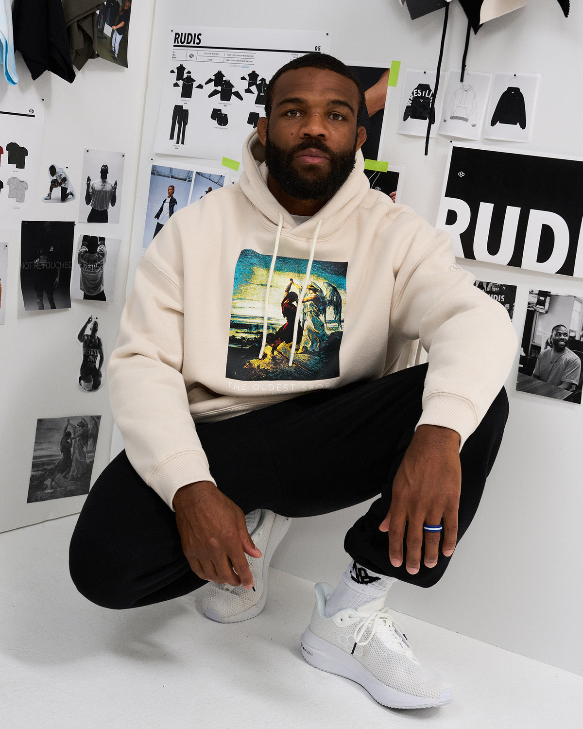 Male model crouching in the RUDIS Divine Struggle fleece hoodie in Bone, showcasing the colorful front artwork, drawstring hood and ribbed cuffs.