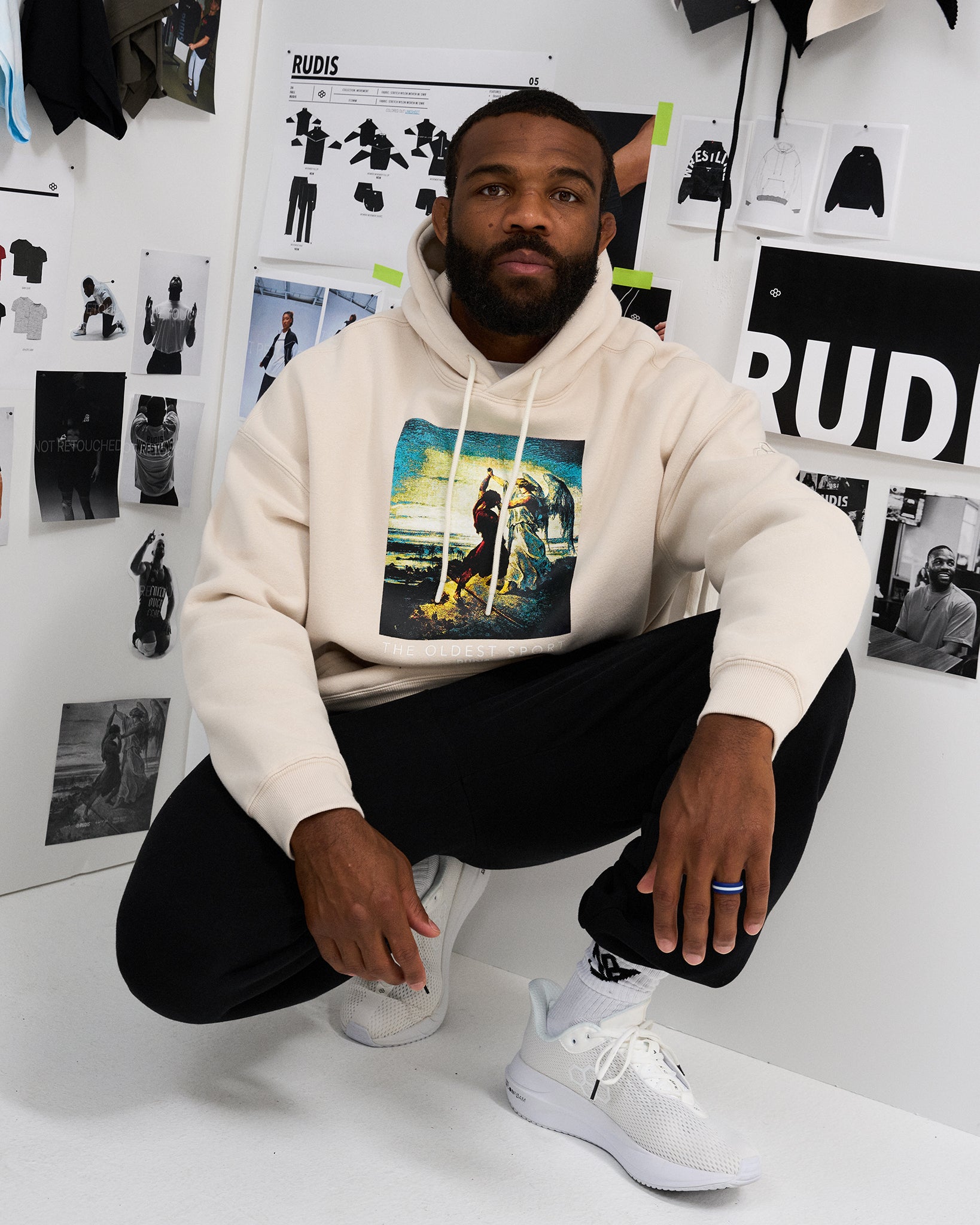 Male model crouching in the RUDIS Divine Struggle fleece hoodie in Bone, showcasing the colorful front artwork, drawstring hood and ribbed cuffs.