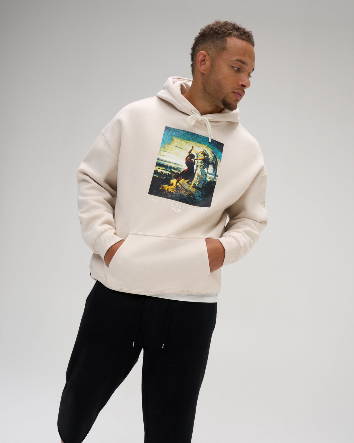 Male model wearing the RUDIS Divine Struggle fleece hoodie in Bone with black pants, showing the relaxed fit, kangaroo pocket and The Oldest Sport graphic.