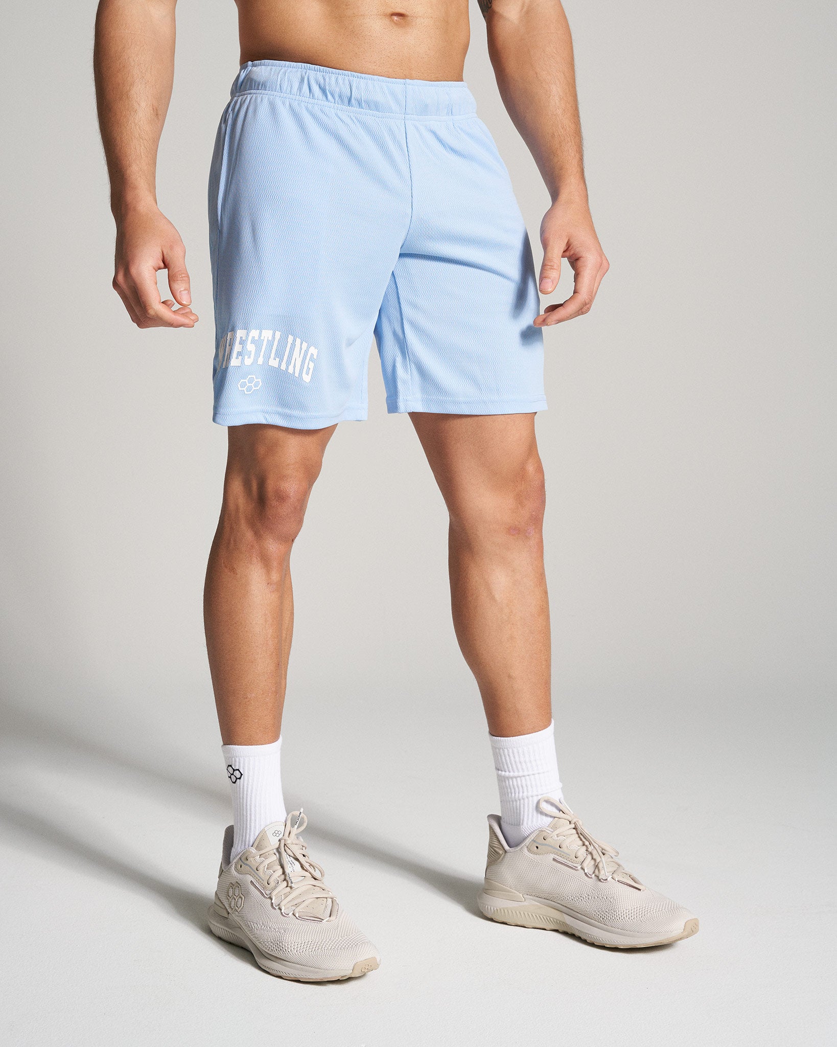 A muscular man is wearing light blue athletic shorts with WRESTLING printed on the side paired with beige running shoes and white socks standing against a neutral background