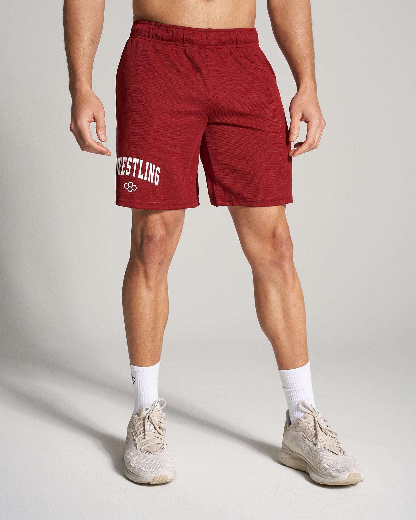 A muscular man is wearing red wrestling shorts showing off an athletic build paired with light beige sneakers and white socks