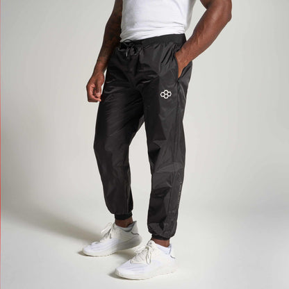 A male model wearing black track pants and white sneakers posing in a relaxed manner against a neutral background