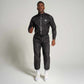 A male model stands confidently in a sleek black athletic outfit featuring a zip-up jacket and matching pants alongside white sneakers against a neutral background