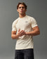 A confident young man stands with a neutral expression wearing a cream-colored t-shirt featuring a subtle logo complemented by dark cargo pants against a muted gray backdrop