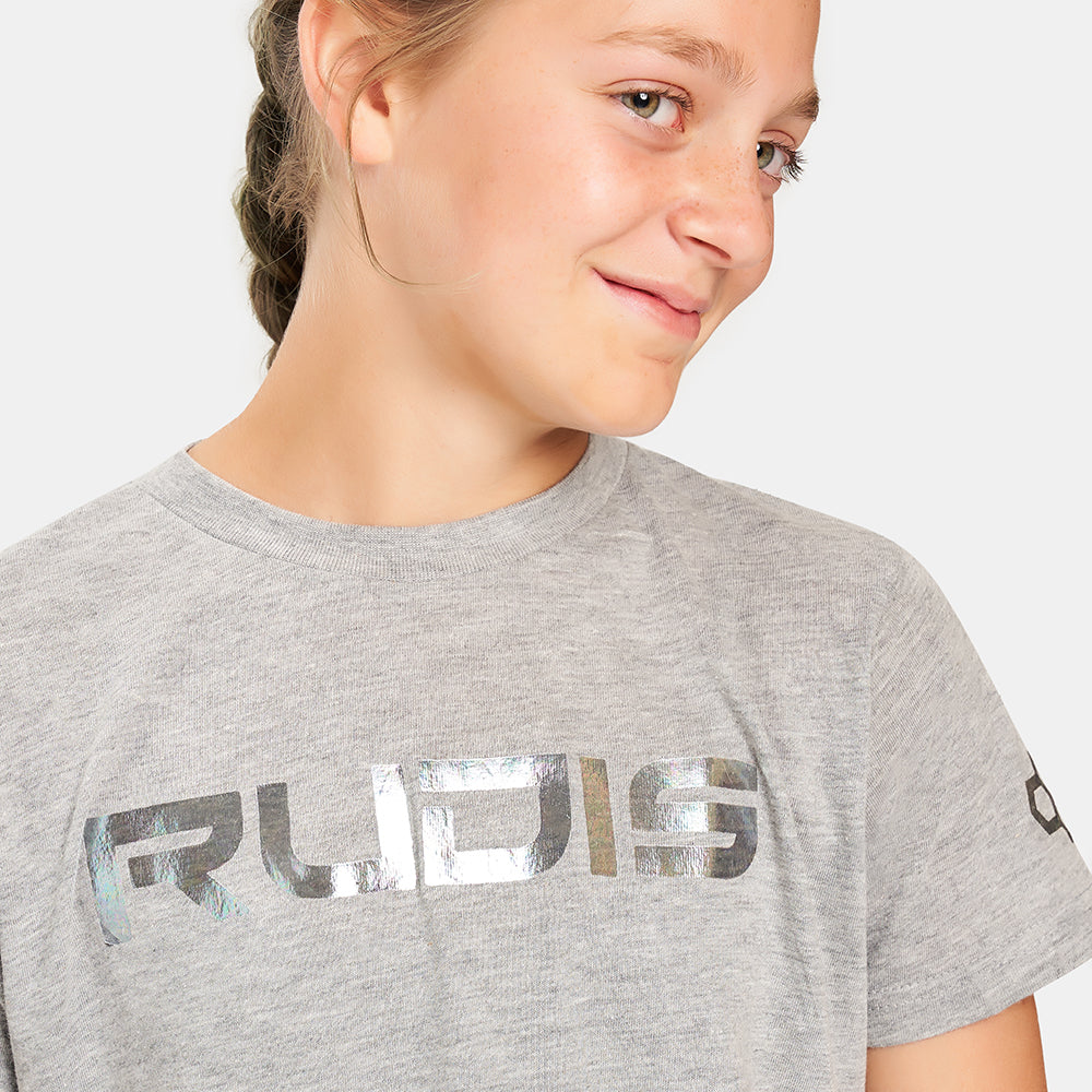 A young girl smiles while wearing a light gray t-shirt featuring a shiny bold logo that reads RUDIS across the front