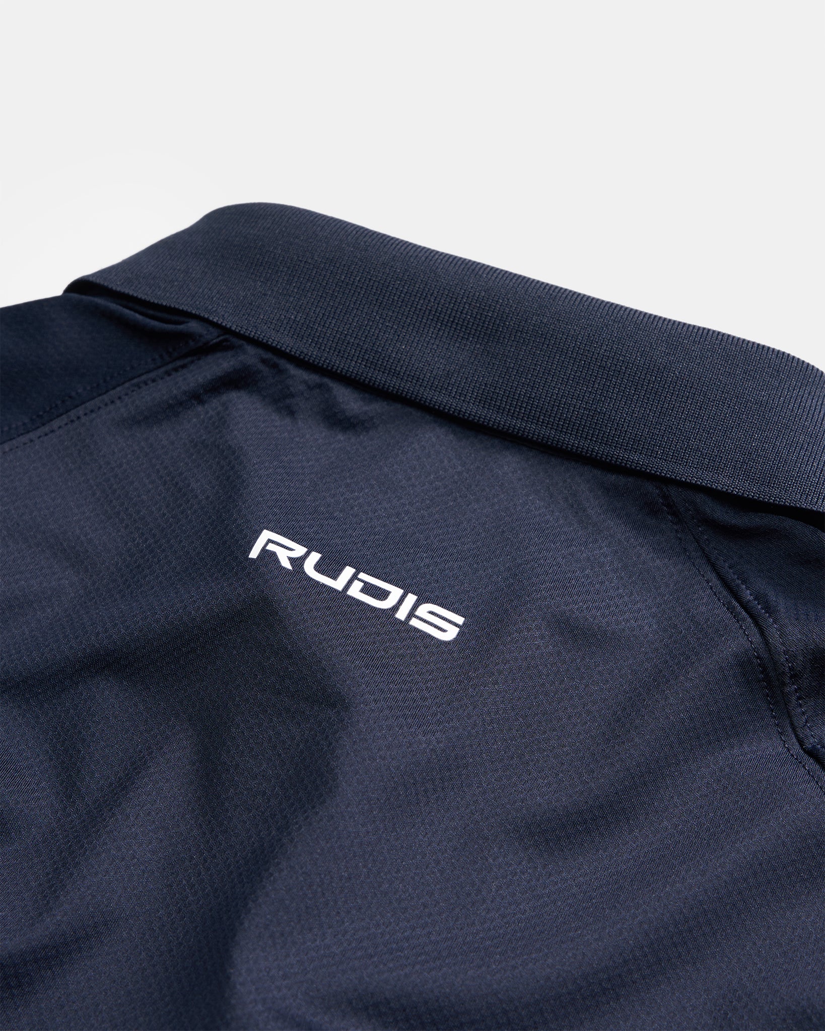 A close-up view of a navy blue sports polo featuring a textured collar and the RUDIS logo embroidered in white