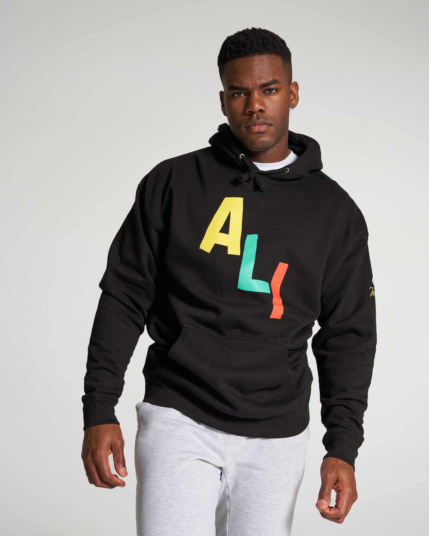 A man is wearing a black hoodie with colorful letters AL! and gray sweatpants posing confidently against a light background