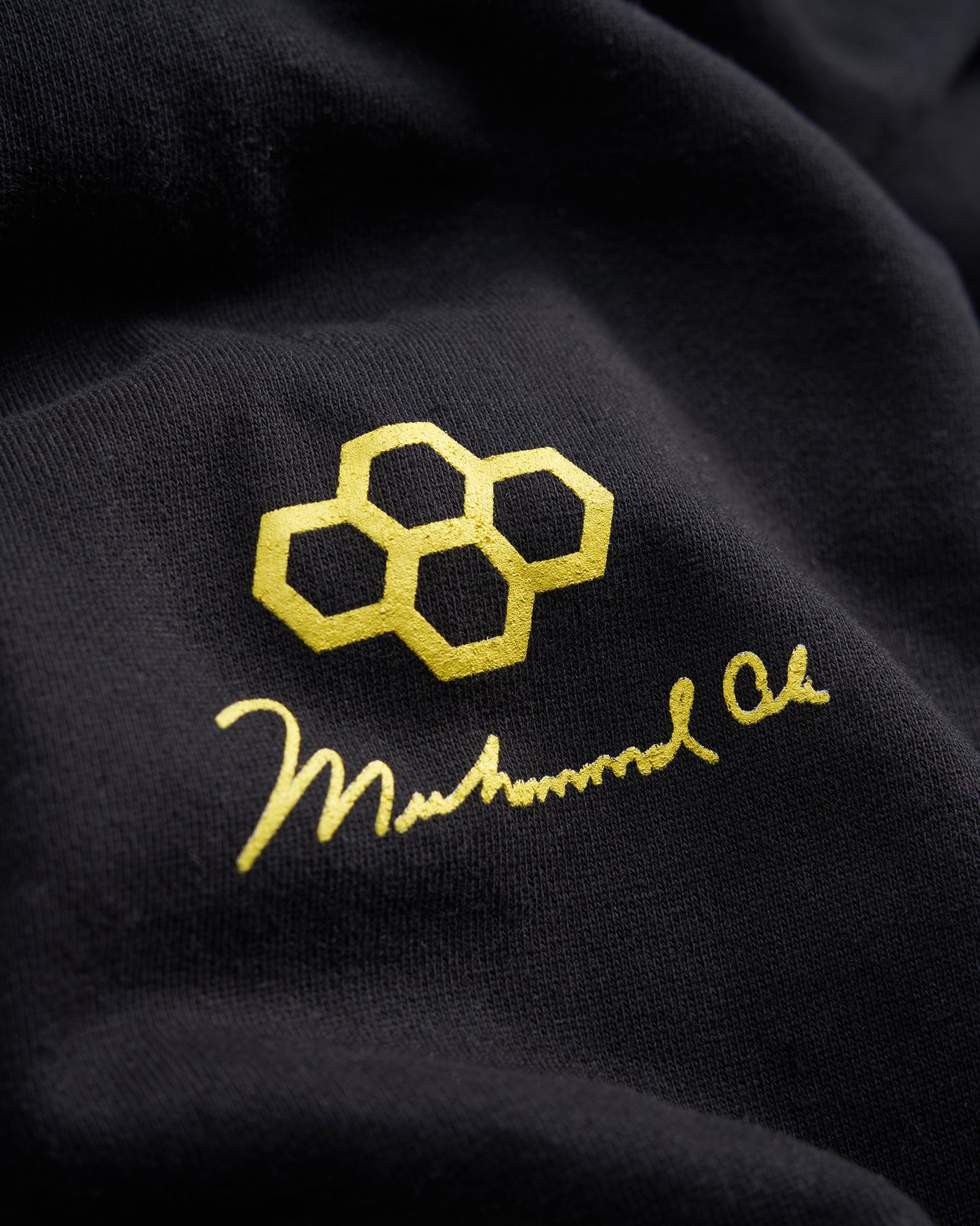A close-up view of a black fabric showcasing a golden logo featuring interconnected hexagons and the signature of Muhammad Ali