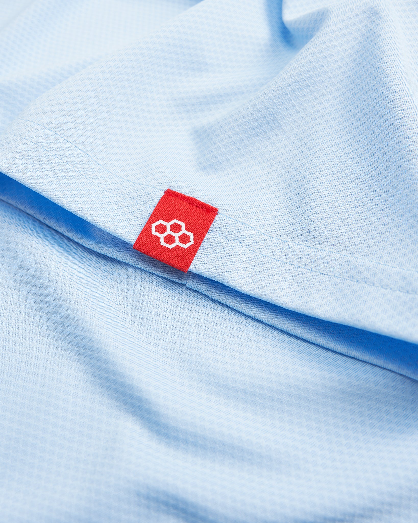 A close-up view of a light blue athletic shirt featuring a textured fabric with a distinctive red label displaying a logo near the hem