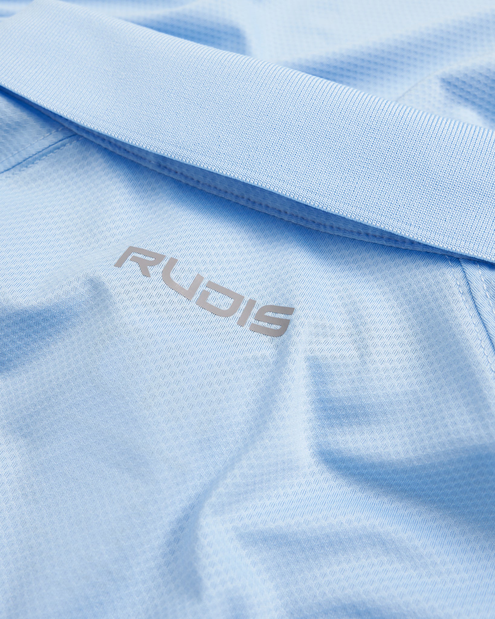 A close-up view of a light blue athletic shirt featuring the RUDIS logo in silver on the fabric