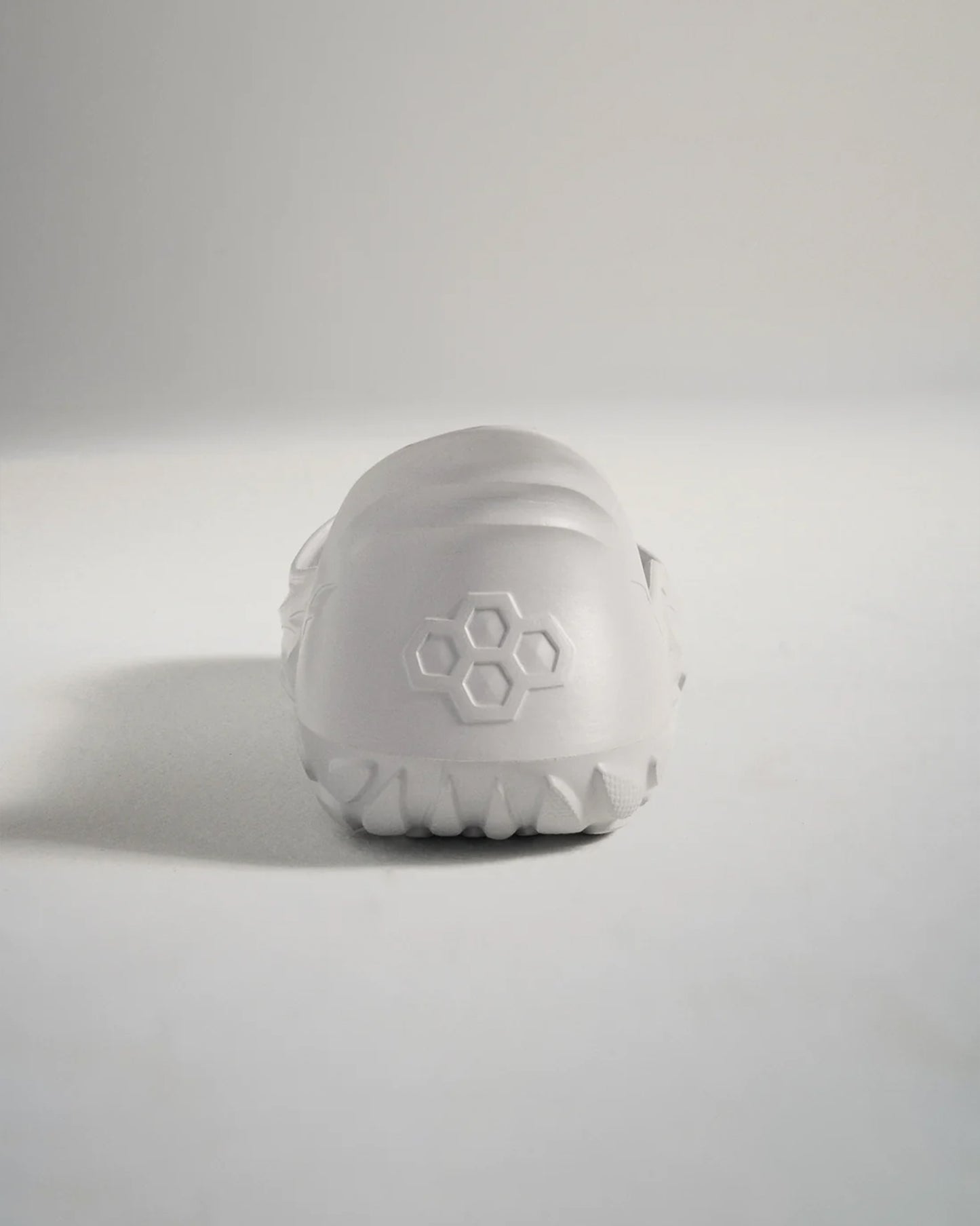 A minimalist white athletic shoe viewed from the back featuring a textured sole and a geometric logo on the heel