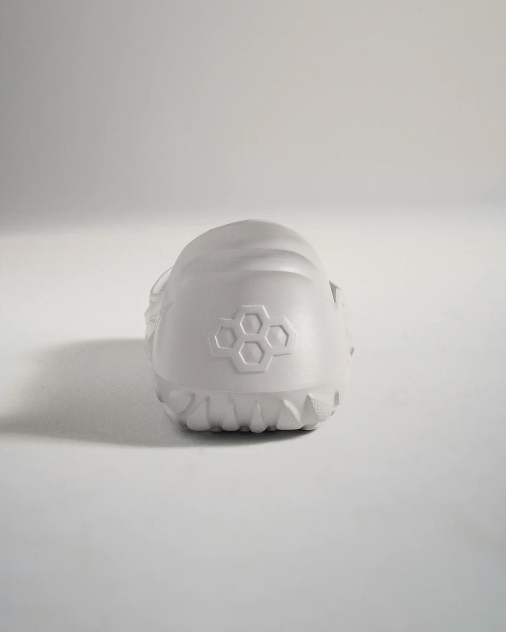 A minimalist white athletic shoe viewed from the back featuring a textured sole and a geometric logo on the heel