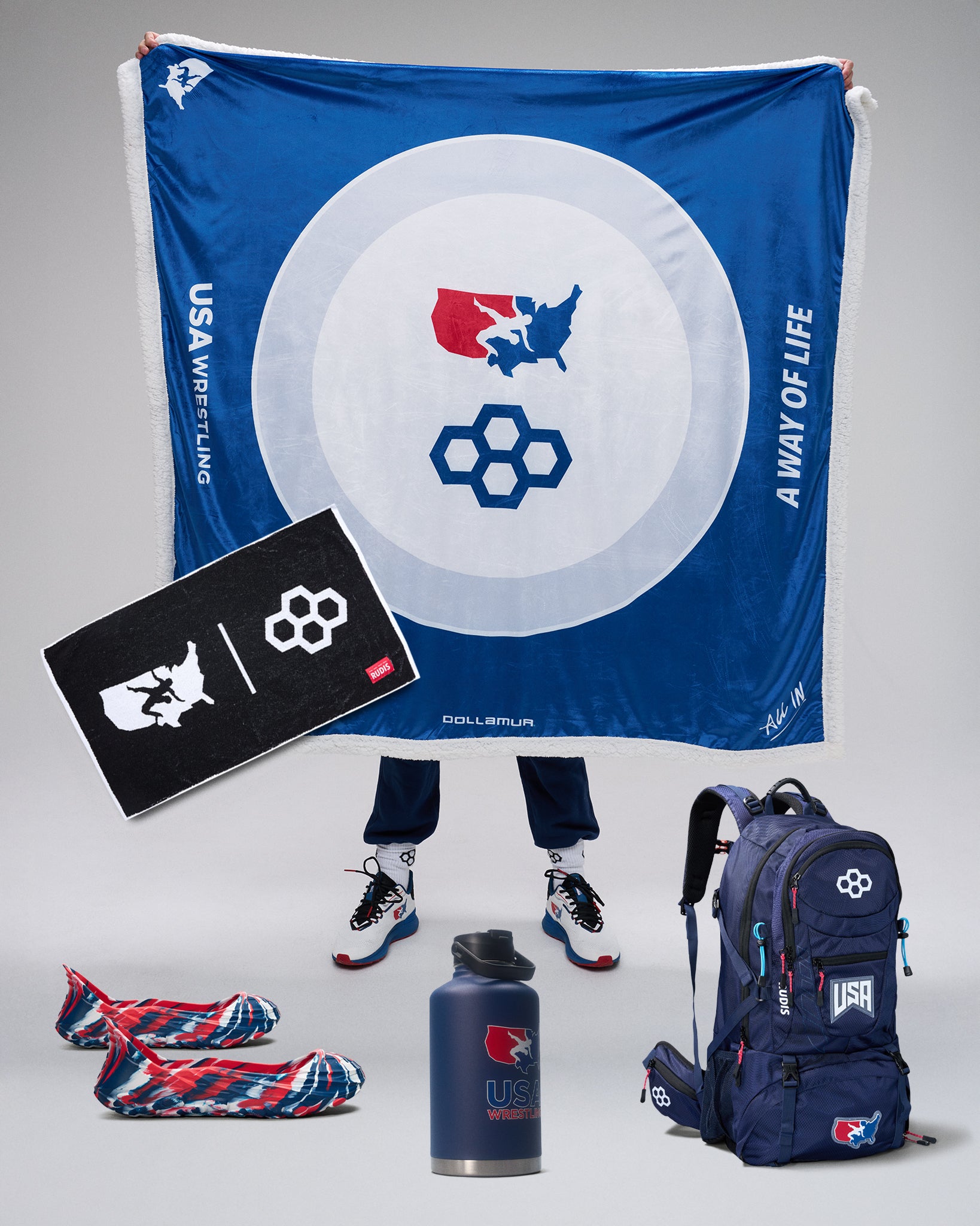 USA Wrestling branded merchandise including a blanket, backpack, towel, and water bottle on a gray background.