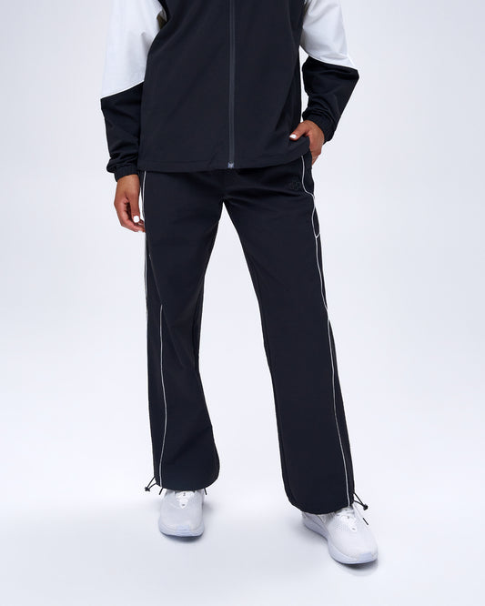 Model wearing black RUDIS Movement Podium Pants with white piping, relaxed legs, and adjustable ankle hems.