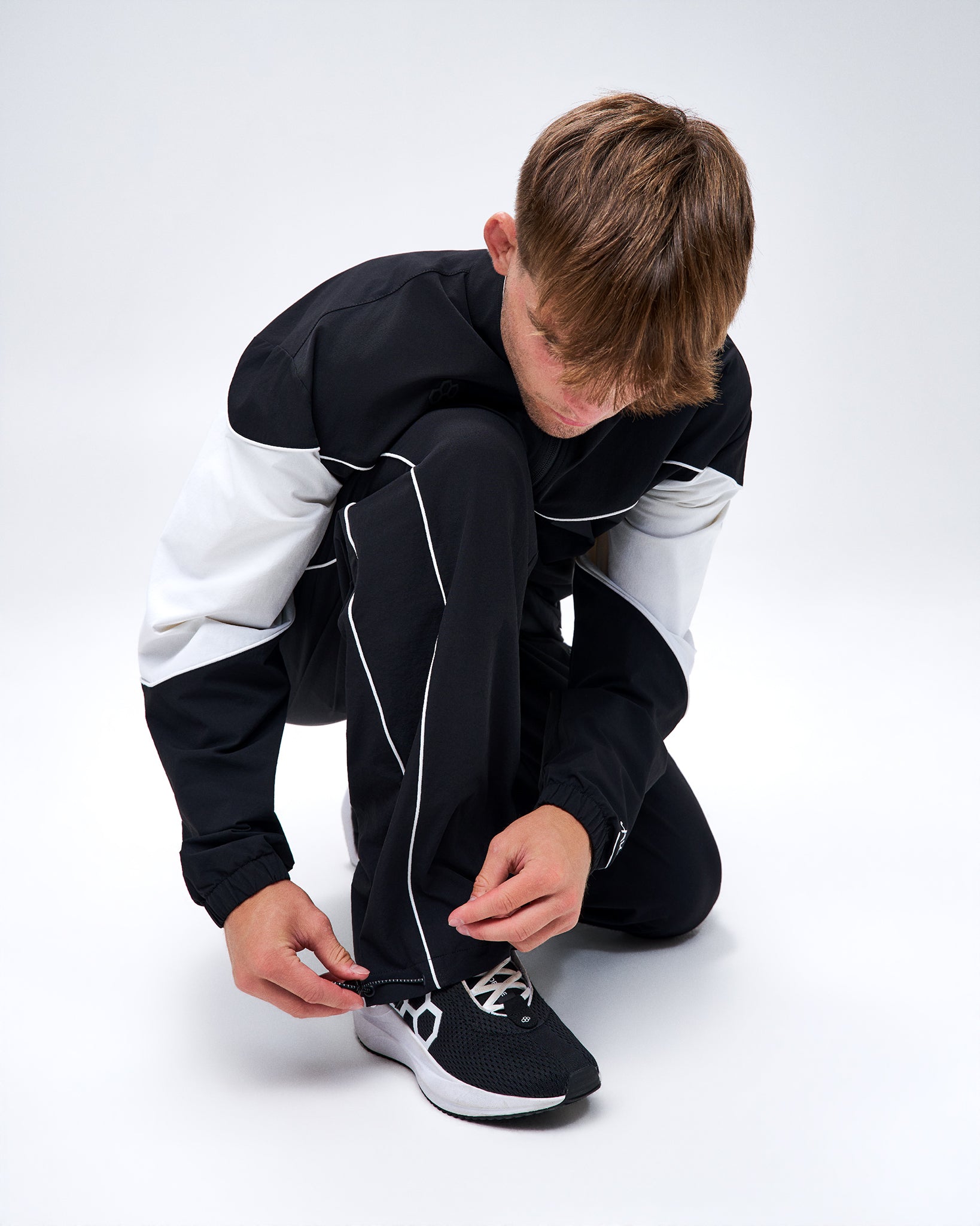 Model adjusting the ankle hem of black RUDIS Movement Podium Pants, highlighting the white piping and adjustable drawcord.