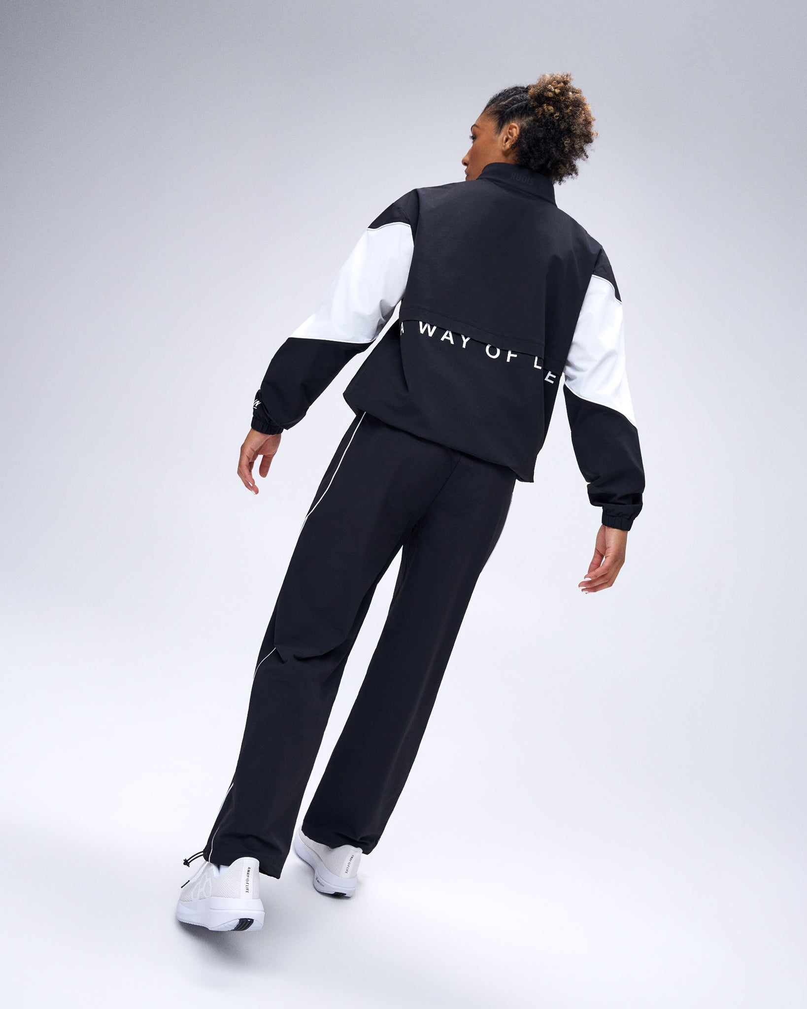 Rear view of model wearing black RUDIS Movement Podium Pants with white piping and matching RUDIS podium jacket.