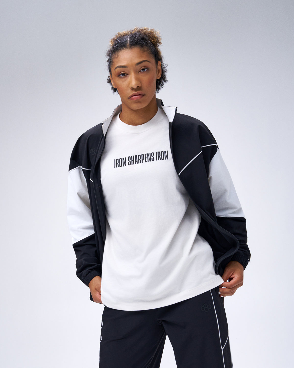 Model wearing the black RUDIS Movement Podium Jacket open over an Iron Sharpens Iron T-shirt with matching black pants.