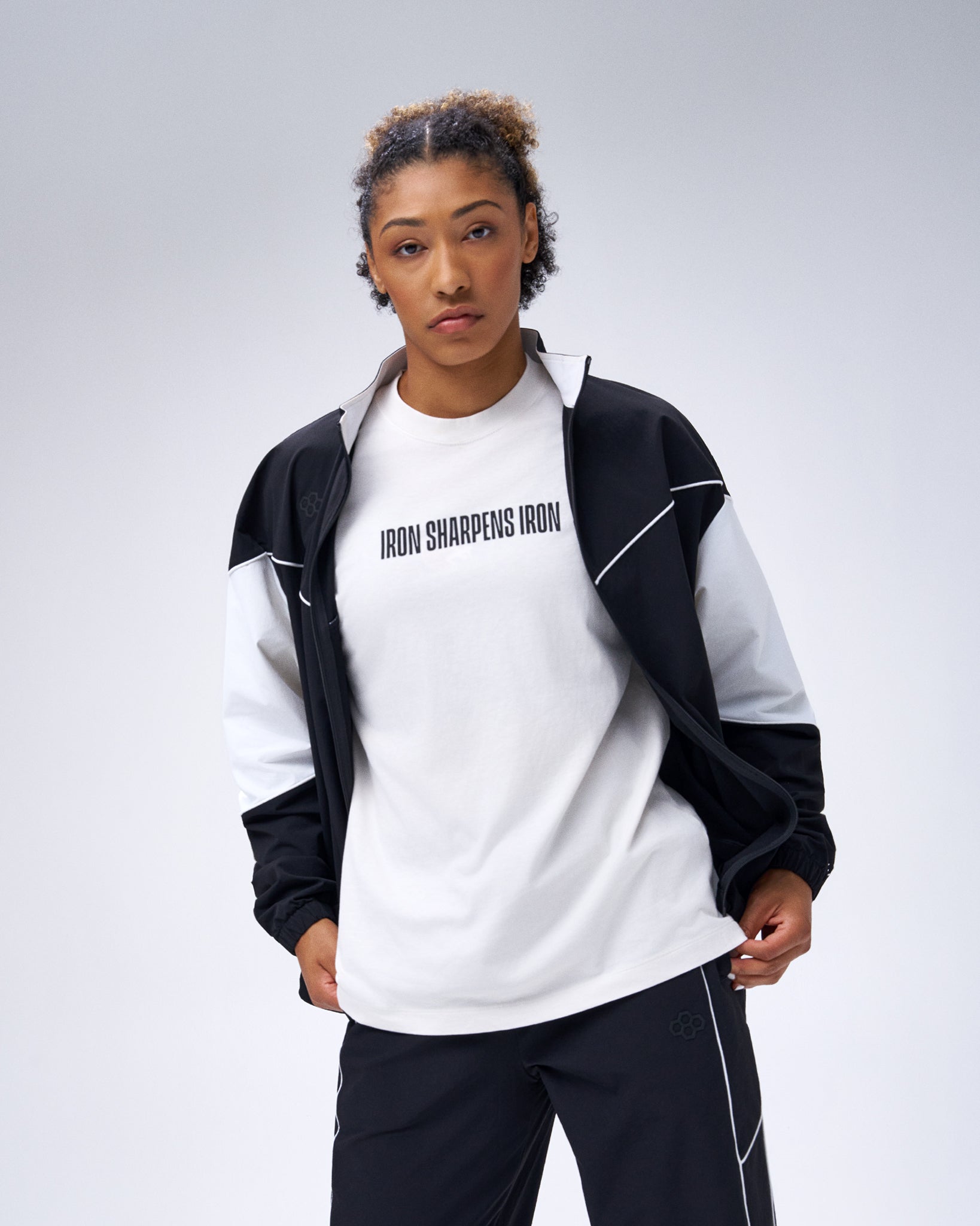 Model wearing the black RUDIS Movement Podium Jacket open over an Iron Sharpens Iron T-shirt with matching black pants.