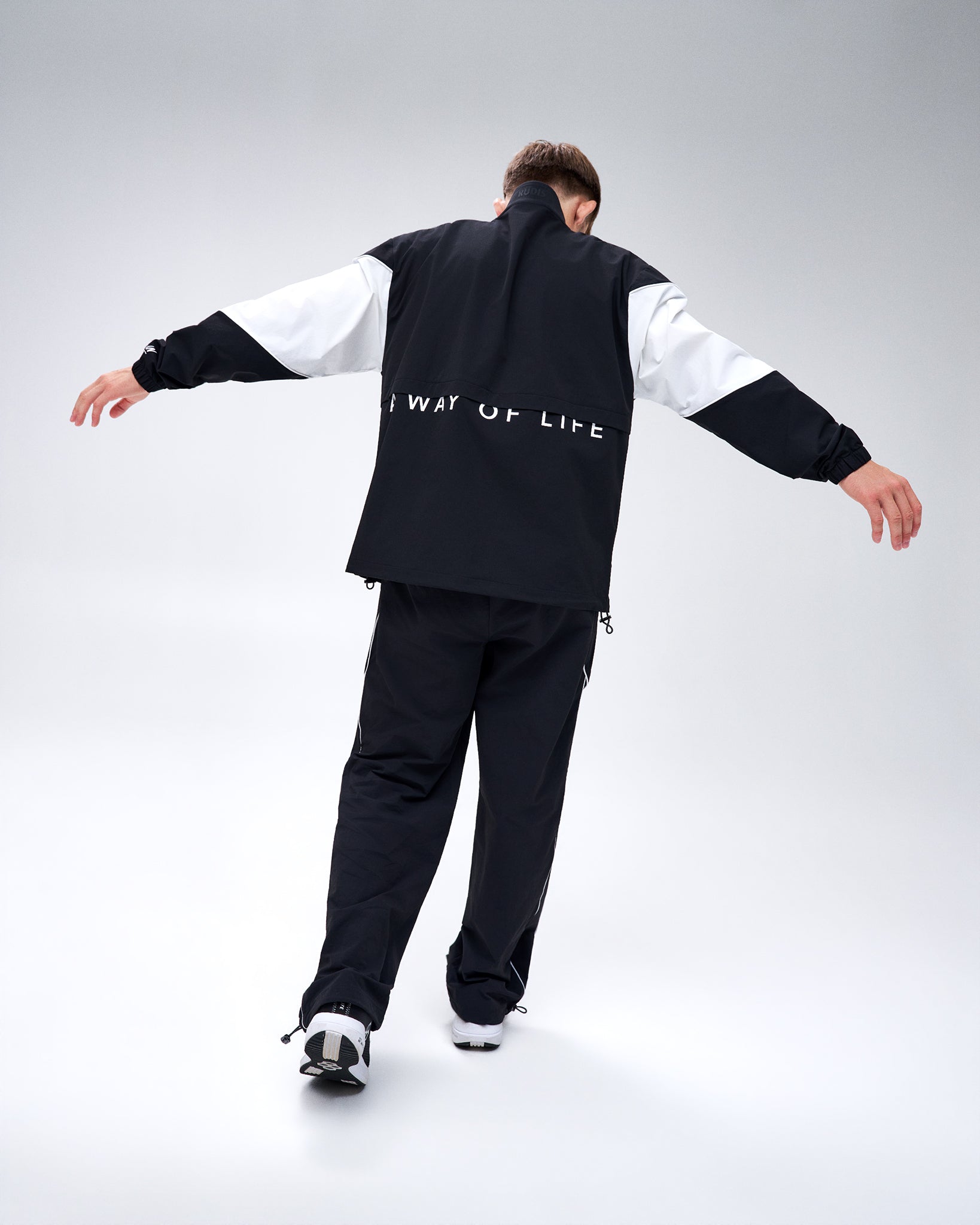 Rear view of model wearing the black RUDIS Full-Zip Movement Podium Jacket and matching pants, showcasing A Way of Life back detailing.