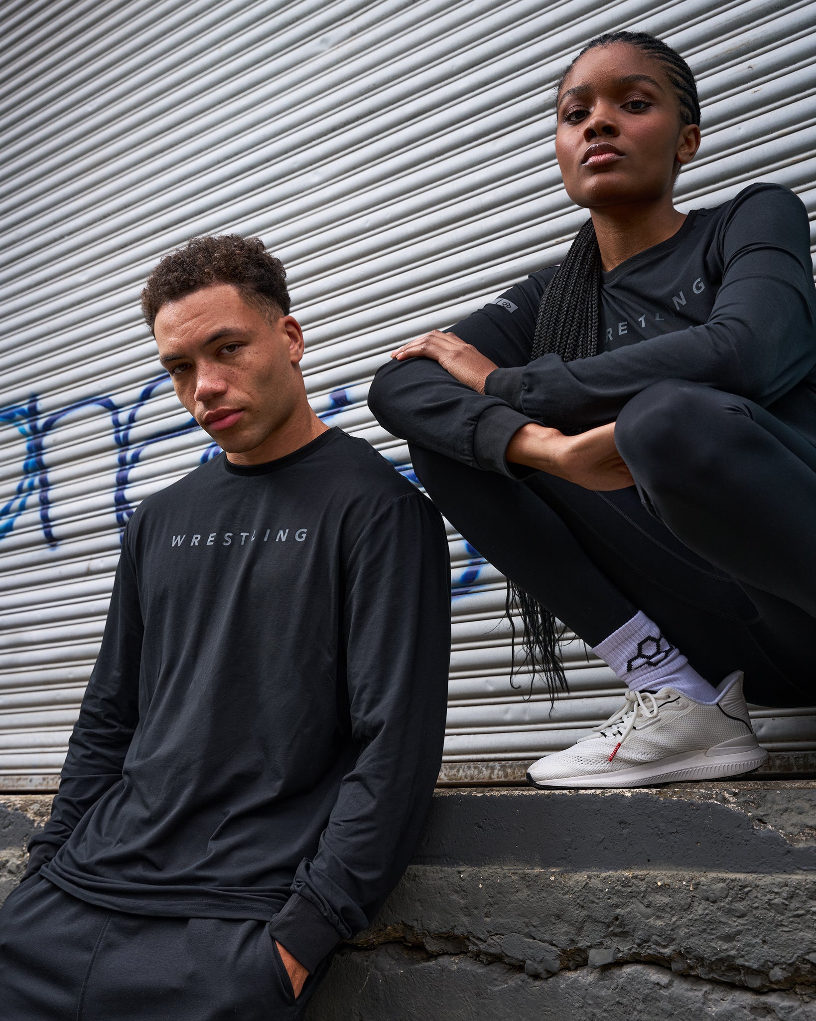 Two people wearing black sweatshirts with visible branding against a metal shutter background.