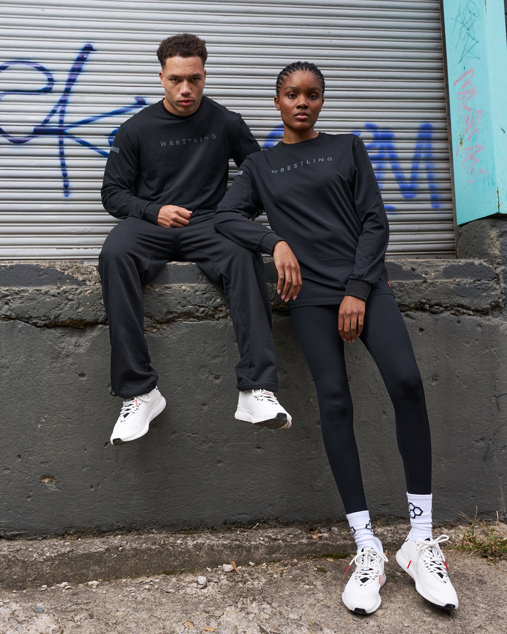 Two people wearing matching black outfits with white sneakers sitting against a graffiti-covered wall.