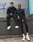 Two people wearing matching black outfits with white sneakers sitting against a graffiti-covered wall.