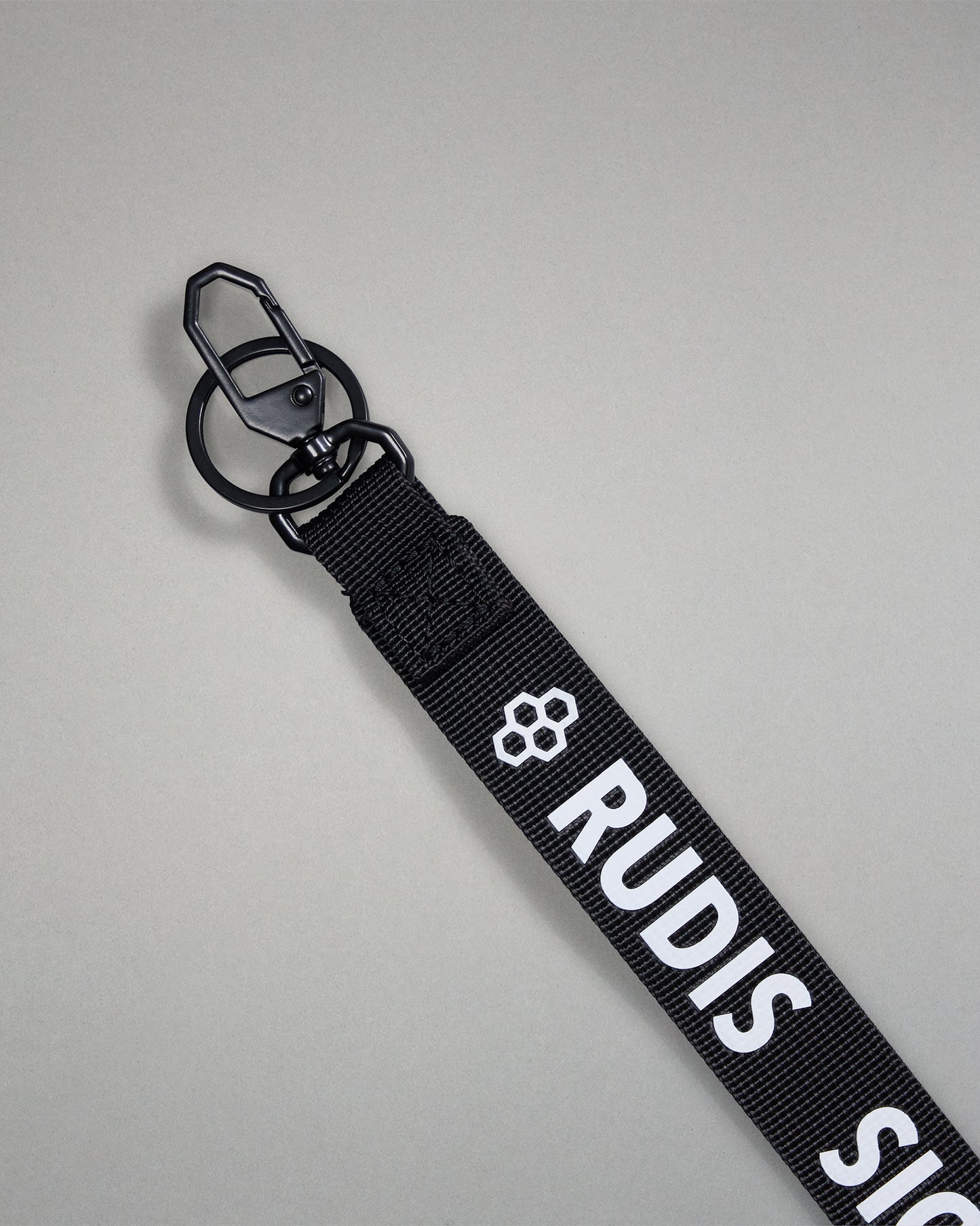 Close-up of the black RUDIS Wordmark lanyard featuring a durable metal key clip, key ring, woven polyester strap, and bold white RUDIS logo with honeycomb icon.