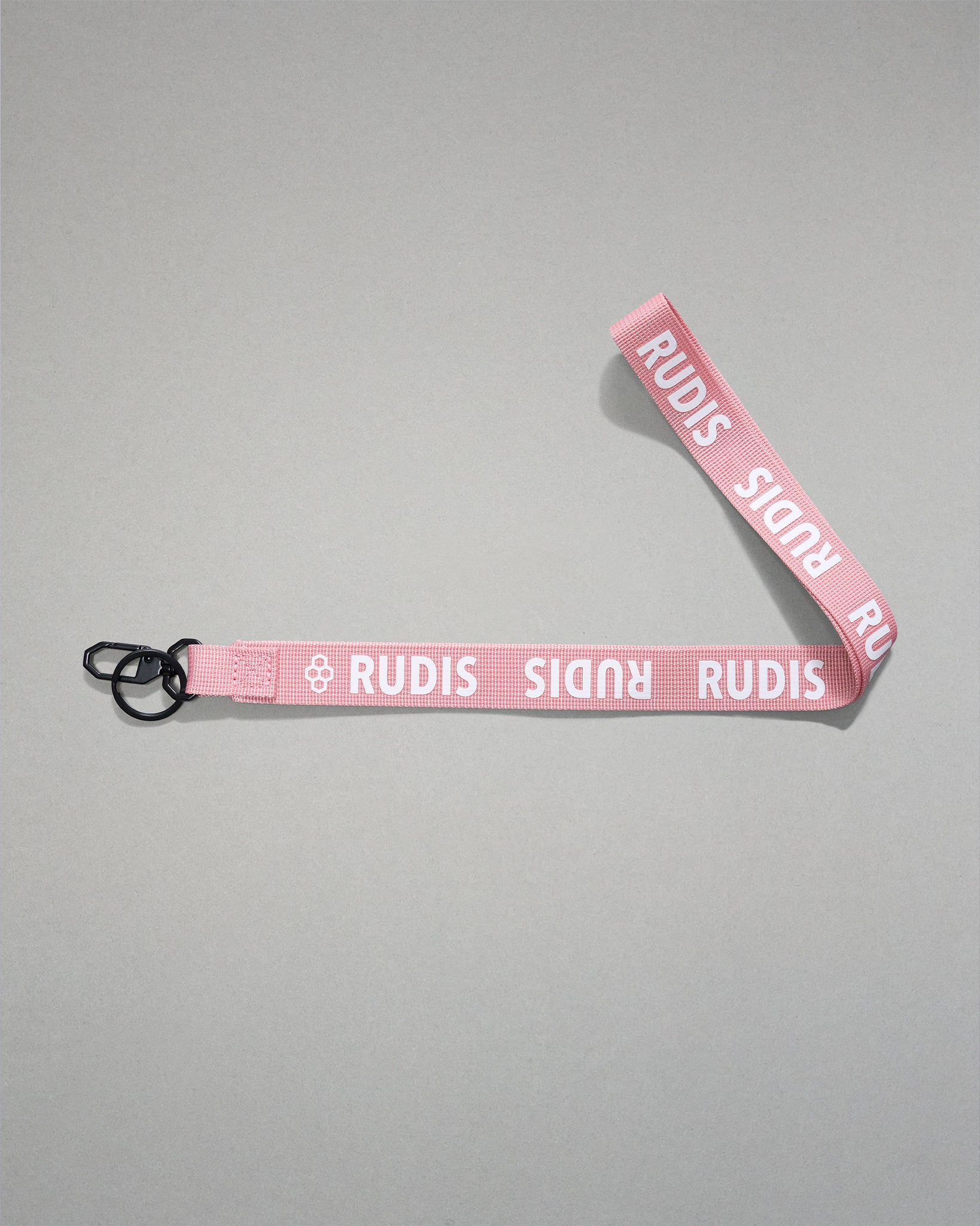 Light pink RUDIS Wordmark lanyard with repeating white RUDIS logos, woven polyester strap, metal key clip, and key ring for carrying keys, badges, or accessories.