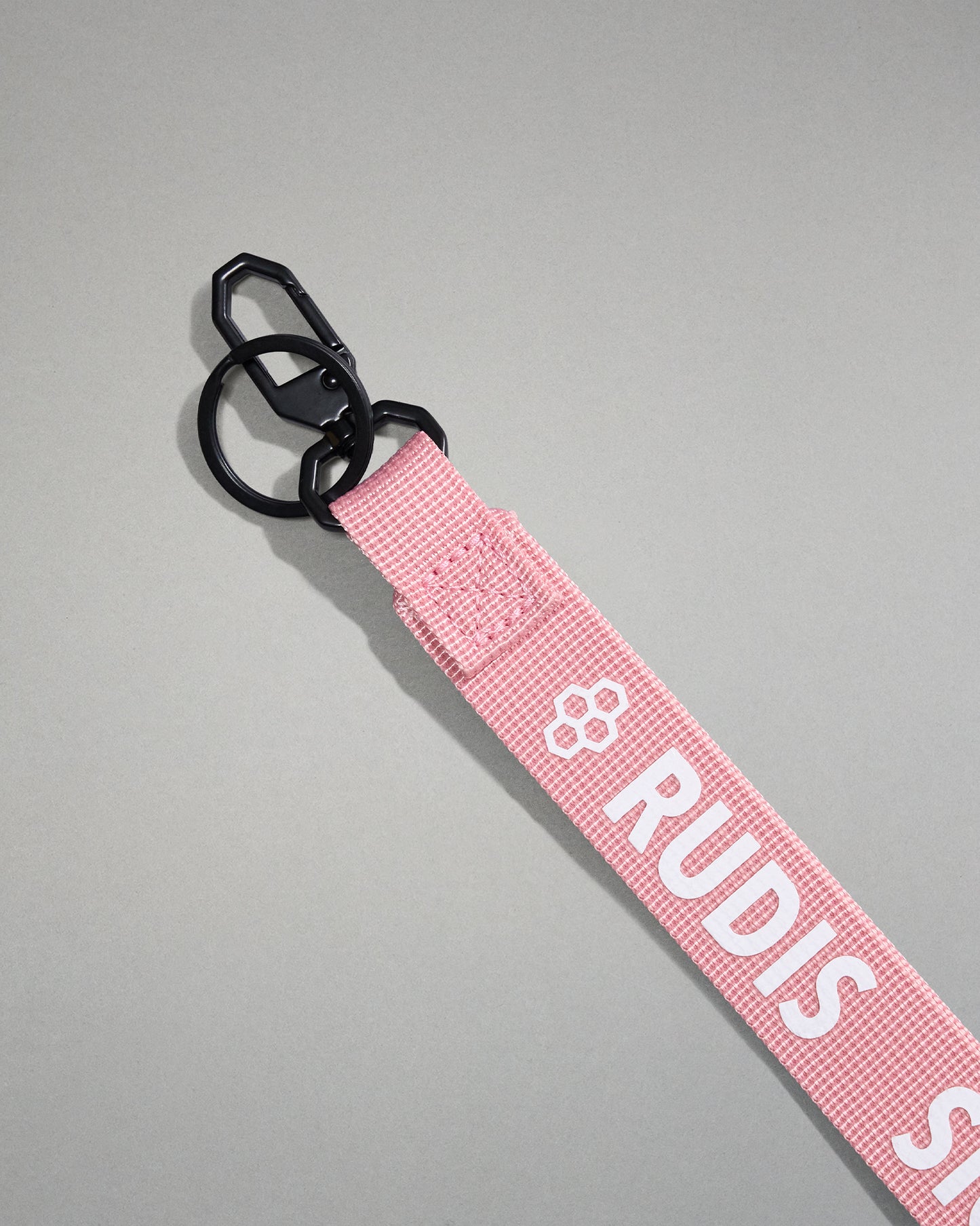 Close-up of the light pink RUDIS Wordmark lanyard featuring a durable metal key clip, key ring, woven polyester strap, and bold white RUDIS logo with honeycomb icon.