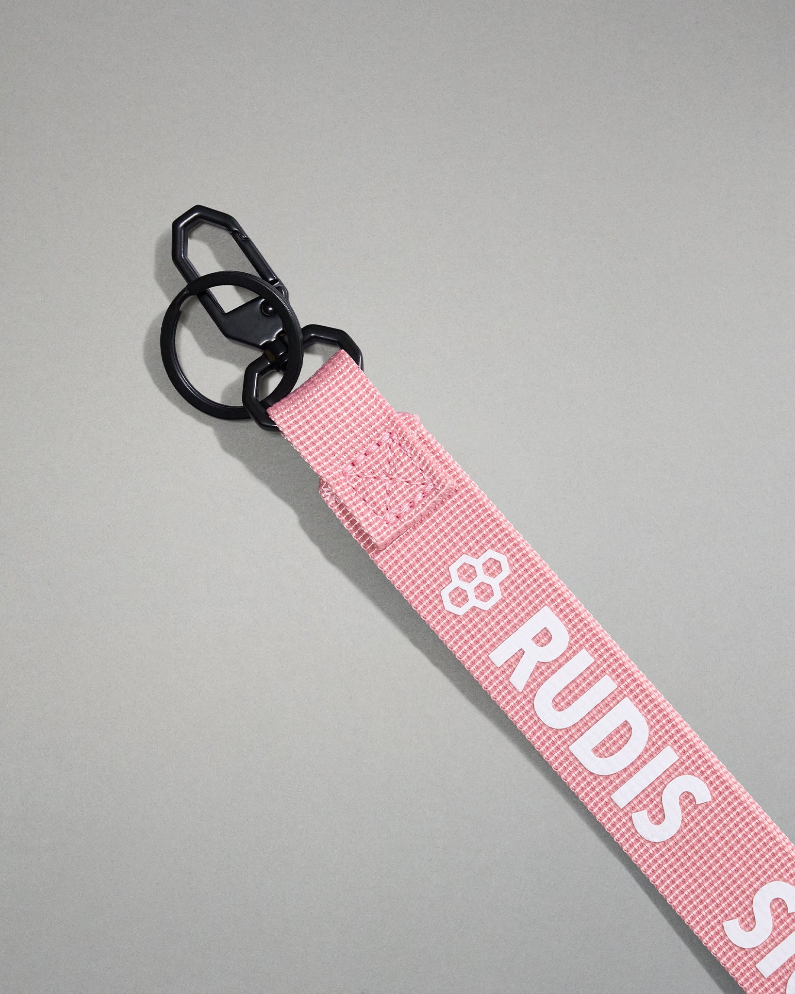 Close-up of the light pink RUDIS Wordmark lanyard featuring a durable metal key clip, key ring, woven polyester strap, and bold white RUDIS logo with honeycomb icon.