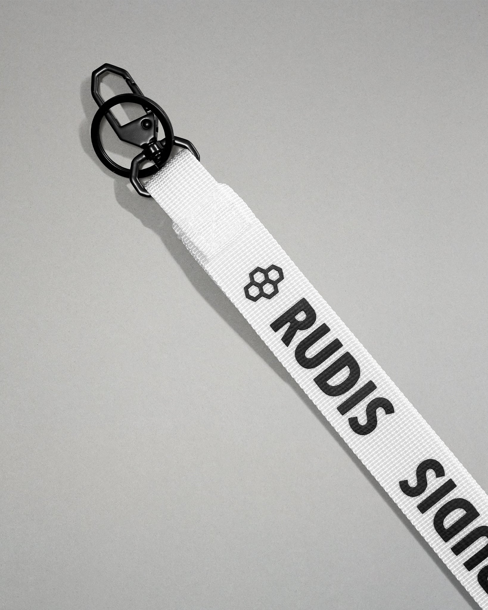 Close-up of the white RUDIS Wordmark lanyard featuring a durable metal key clip, key ring, woven polyester strap, and bold white RUDIS logo with honeycomb icon.