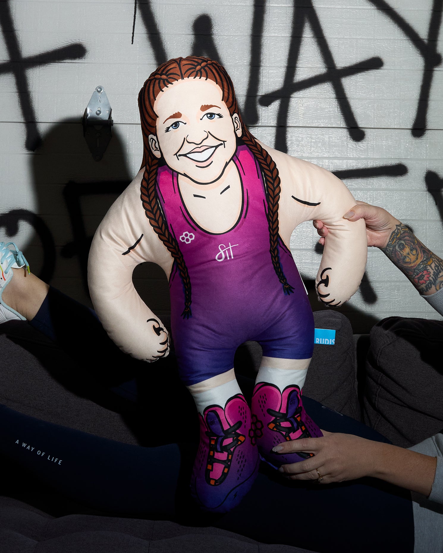 Pillow of a person in a purple singlet with tattoos, held by a person against a graffiti-covered wall.