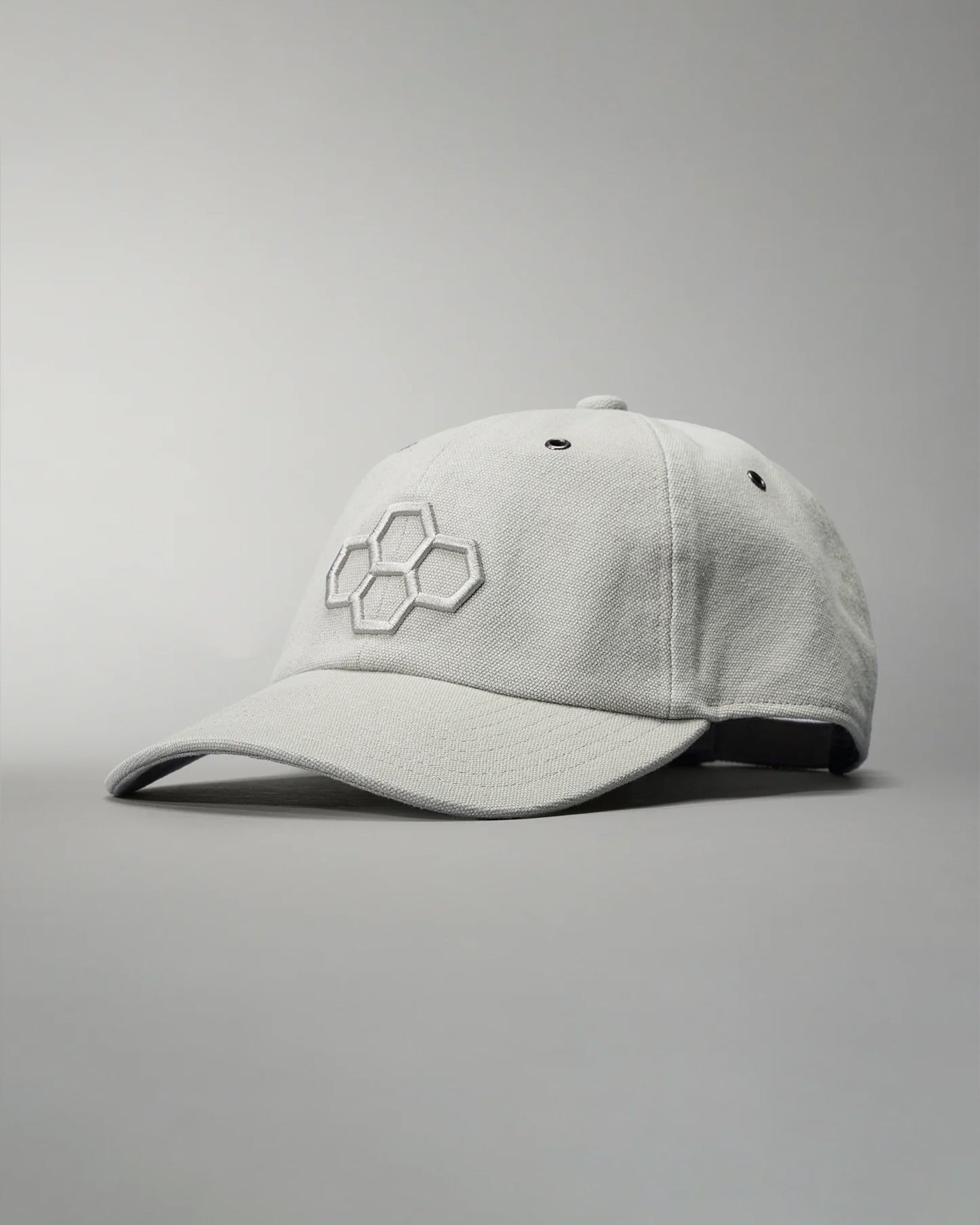 A light gray baseball cap with an embroidered hexagonal logo is displayed at an angle against a muted gray background, highlighting its structured design and comfortable fabric.