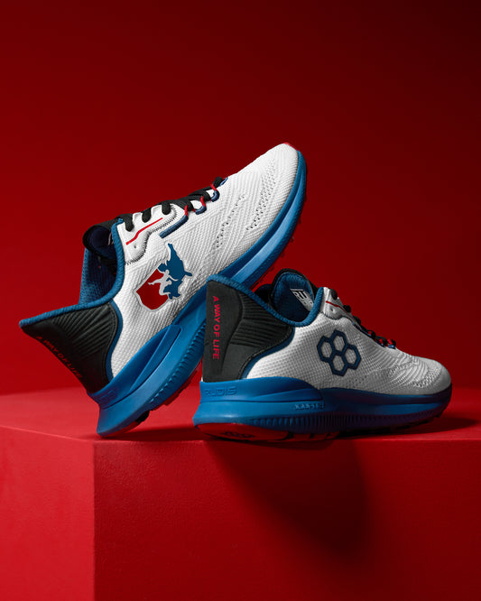 Side view of a pair of white RUDIS training shoes with blue soles, black and red accents, and USA Wrestling and hexagon logos. One heel features the phrase ‘A Way of Life’ in red text. Shoes are displayed on a red background.
