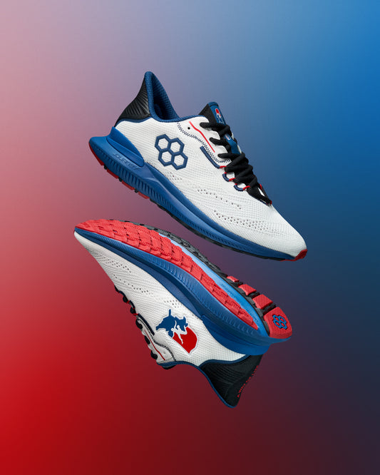 Pair of white RUDIS training shoes with blue midsoles, red and black detailing, and hexagon and USA Wrestling logos. The outsoles feature a red tread pattern. Shoes are displayed floating against a red-to-blue gradient background.