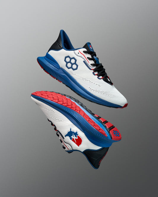 Pair of white RUDIS training shoes with blue midsoles, red and black detailing, and hexagon and USA Wrestling logos. The outsoles feature a red tread pattern. Shoes are displayed floating against a red-to-blue gradient background.