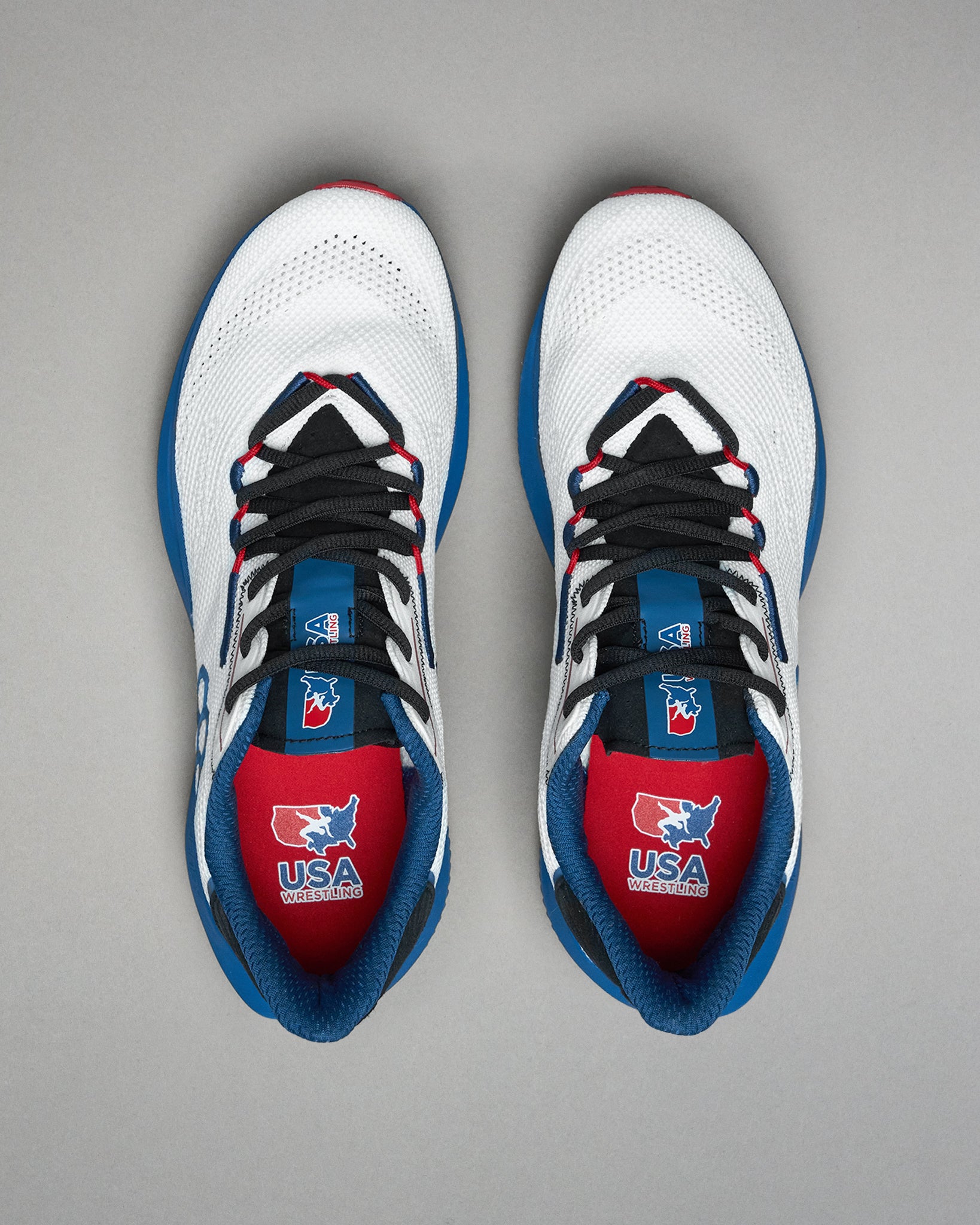 Top view of a pair of white RUDIS training shoes with black laces, blue and red accents, and USA Wrestling logos visible on the red insoles and blue tongues. Displayed on a light gray background.