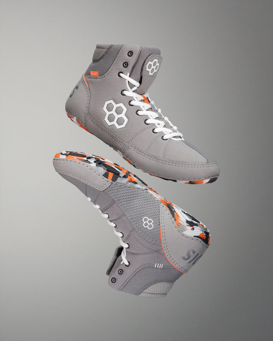 High-top wrestling shoes feature a gray upper with vibrant orange and black accents, designed for style and support, showcased against a soft gradient background.