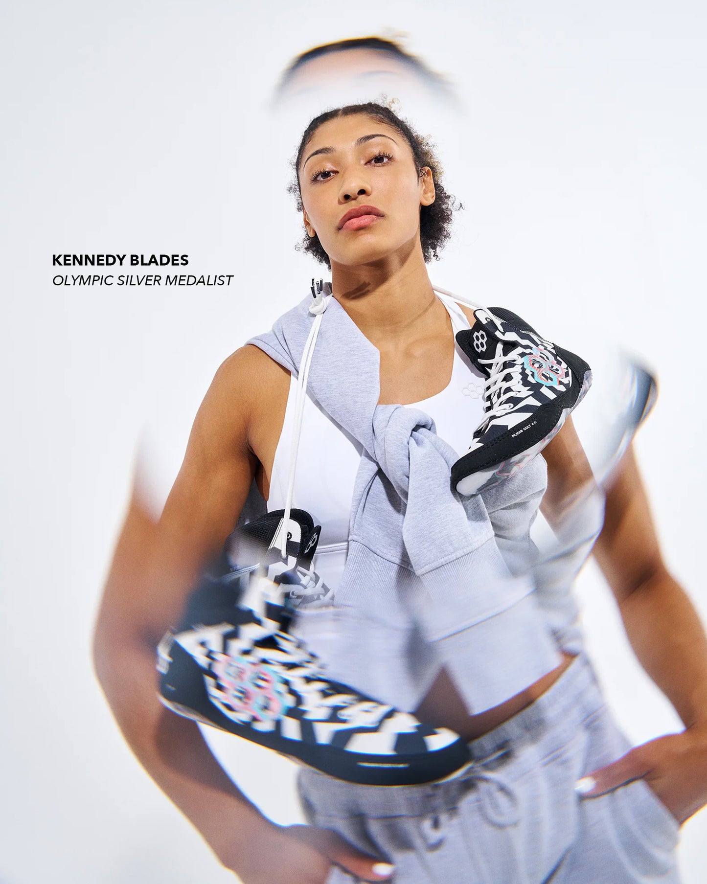 A woman in a light gray athletic outfit holds up a black and white patterned sneaker, with a second identical sneaker blurred in front of her. Text on the left reads 'KENNEDY BLADES OLYMPIC SILVER MEDALIST'.