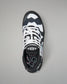 A single athletic wrestling shoe shown from a top-down view on a neutral gray background. The shoe features a black and white geometric pattern with white laces and a black tongue displaying a white logo.