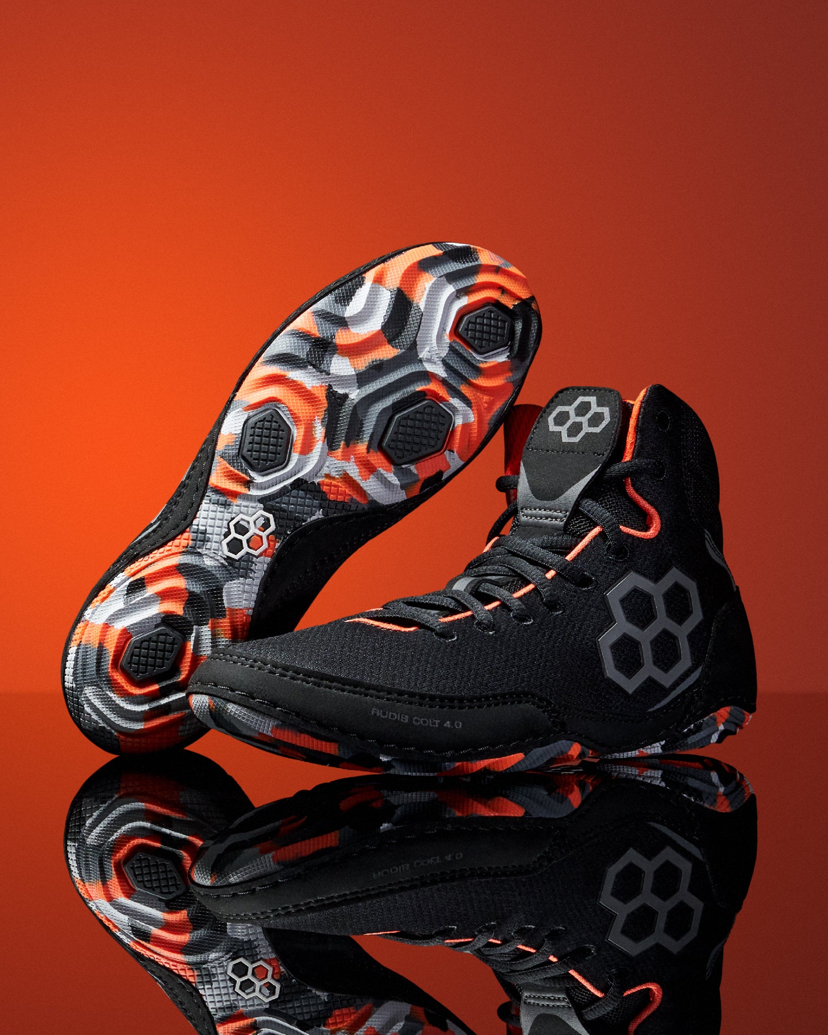 A pair of black Wrestling shoes with vibrant orange and gray geometric tread patterns, displayed on a reflective surface against a solid orange background.