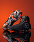 A pair of black Wrestling shoes with vibrant orange and gray geometric tread patterns, displayed on a reflective surface against a solid orange background.