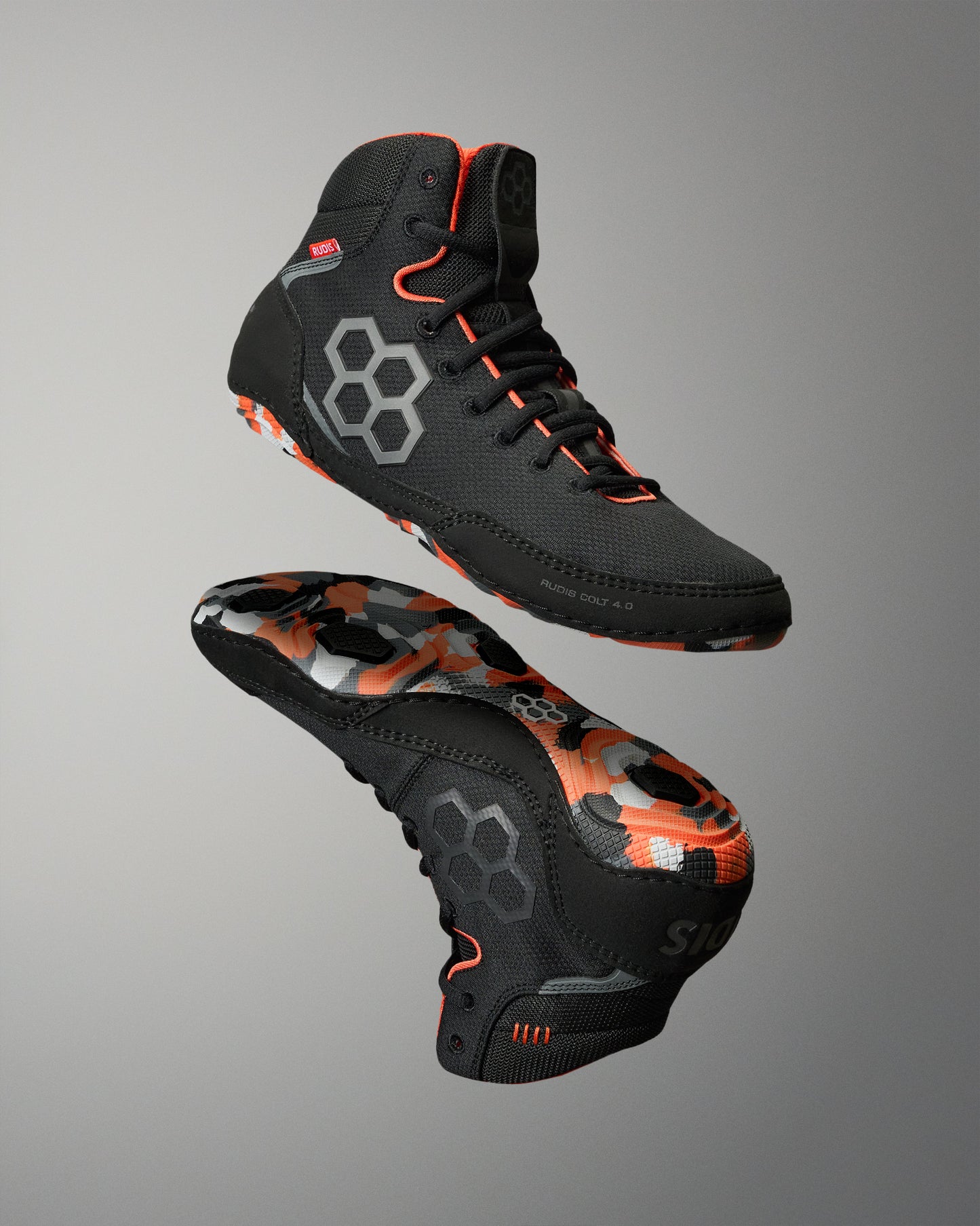 A pair of high-top Wrestling shoes shown floating against a neutral gray gradient background, displaying both the black upper with gray hexagonal accents and the black sole with a vibrant orange and gray camouflage pattern.