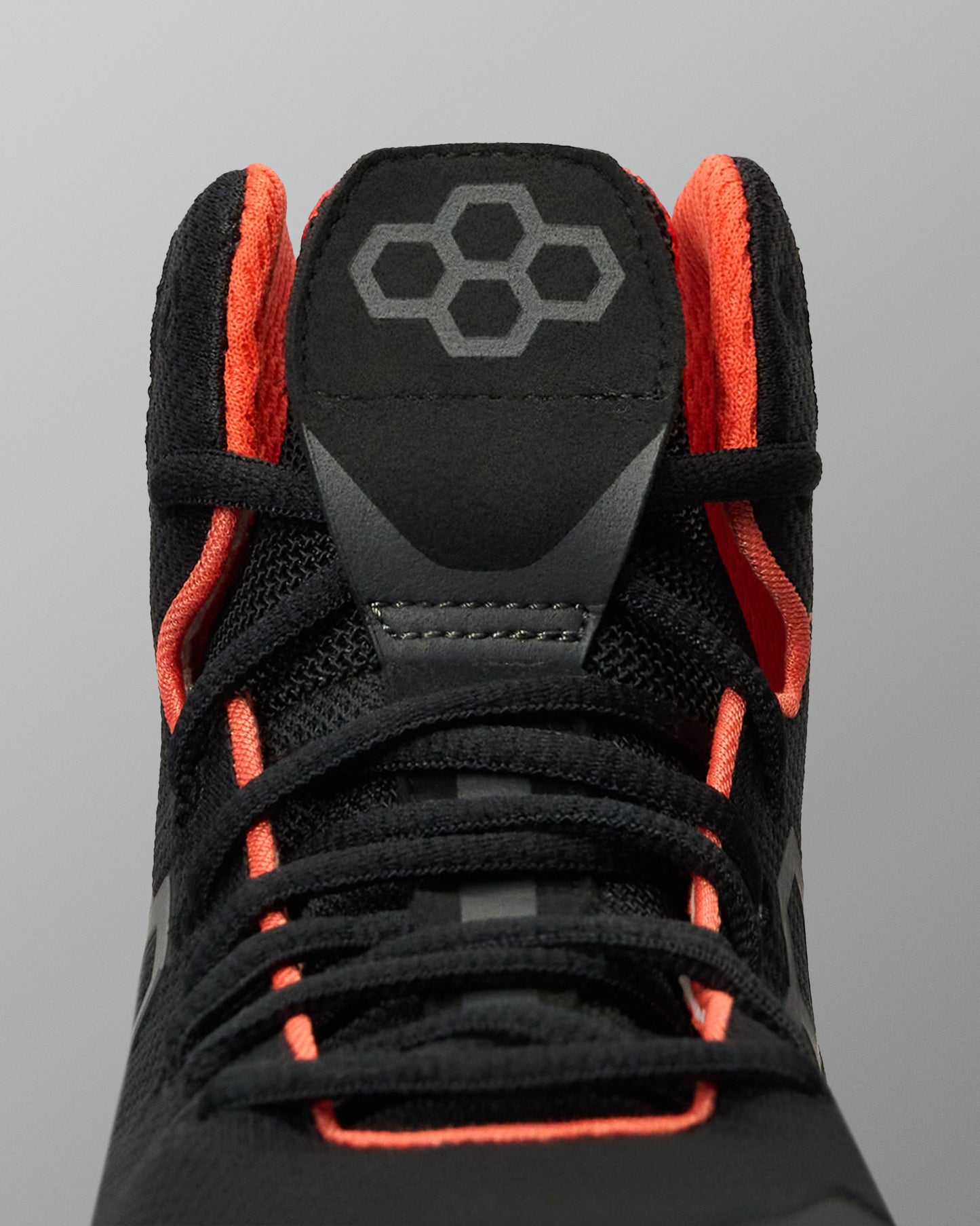 Close-up of a black Wrestling shoe with bright orange lining and laces, featuring a gray geometric logo on the tongue, set against a solid gray background.