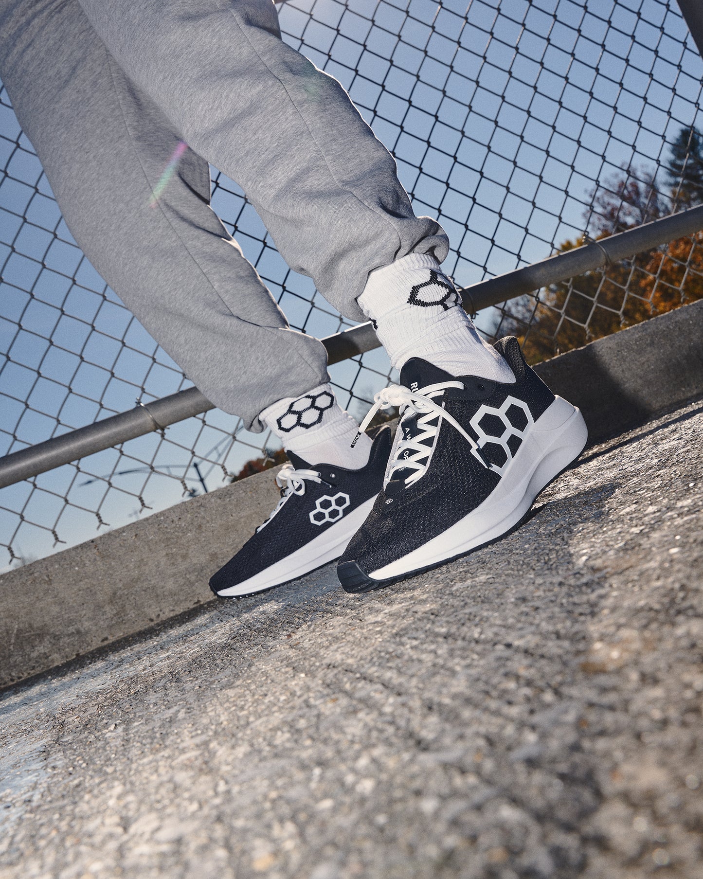 Person wearing black and white sneakers with geometric patterns, standing on a concrete surface with a metal railing and chain-link fence in the background.