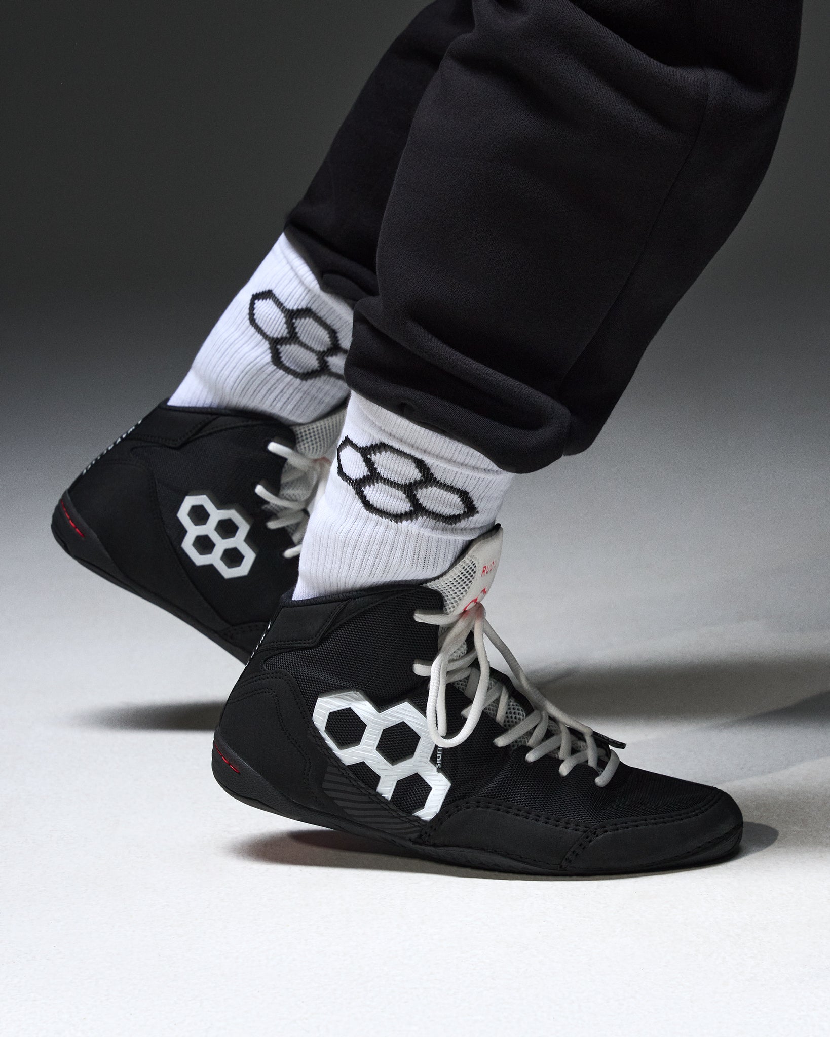 Black high-top wrestling shoes with white geometric hexagonal logos, worn with white crew socks featuring black logos and black sweatpants, shown against a dark gray studio background.
