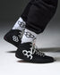 Black high-top wrestling shoes with white geometric hexagonal logos, worn with white crew socks featuring black logos and black sweatpants, shown against a dark gray studio background.